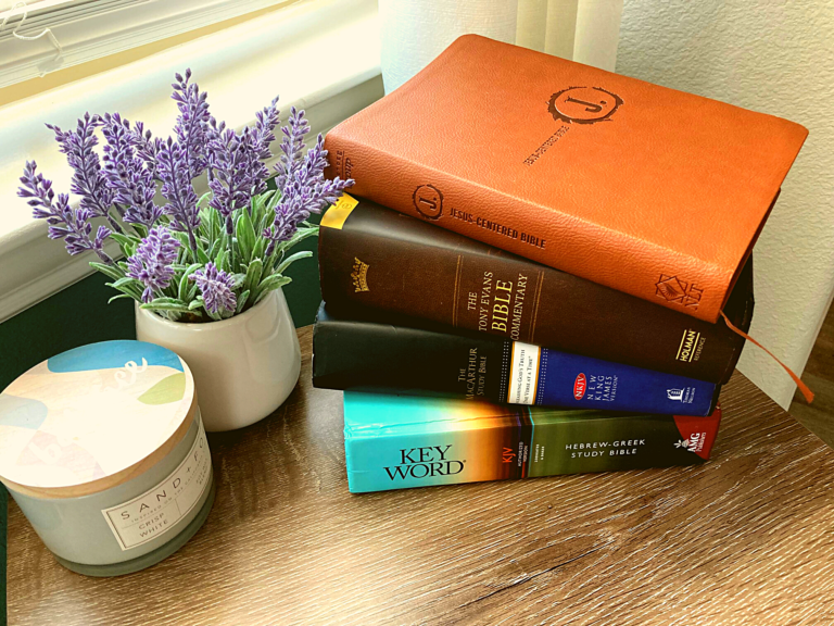 Stack of Bibles on table next to purple flowers and candle.