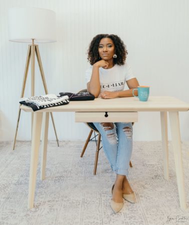 African American Christian entrepreneur woman in white t-shirt sitting at desk with t-shirts and coffee mug on desk.