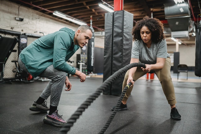 Woman doing battle ropes in gym while male trainer watches