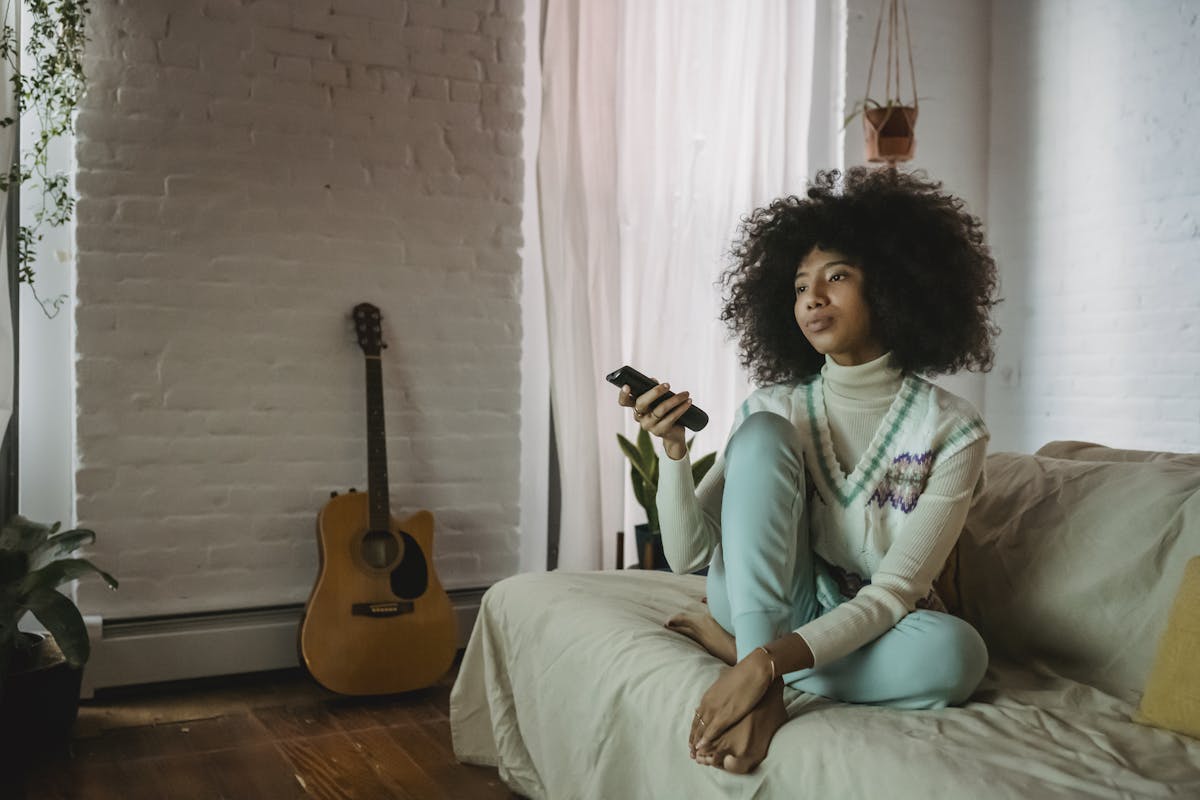 Young woman with curly hair relaxing on a couch with a remote control in a cozy living room.