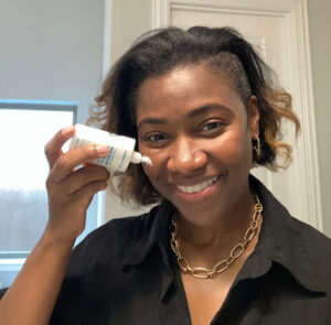 Black woman smiling in mirror as she holds a bottle of sunscreen on her right cheek. She is in a bathroom and wearing a black shirt with gold earrings and a old chain necklace.
