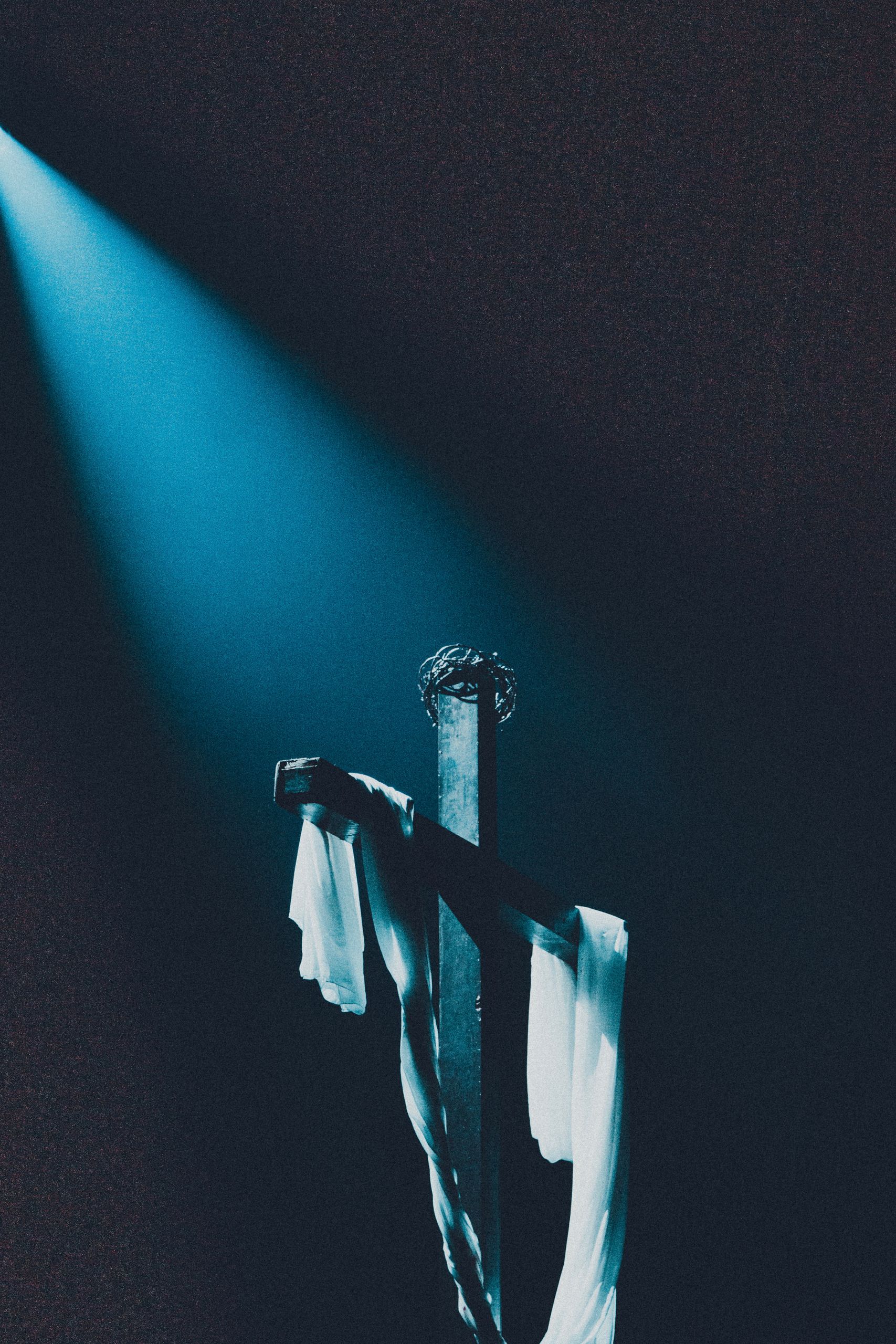A wooden cross with white fabric under a dramatic spotlight in a dark room.
